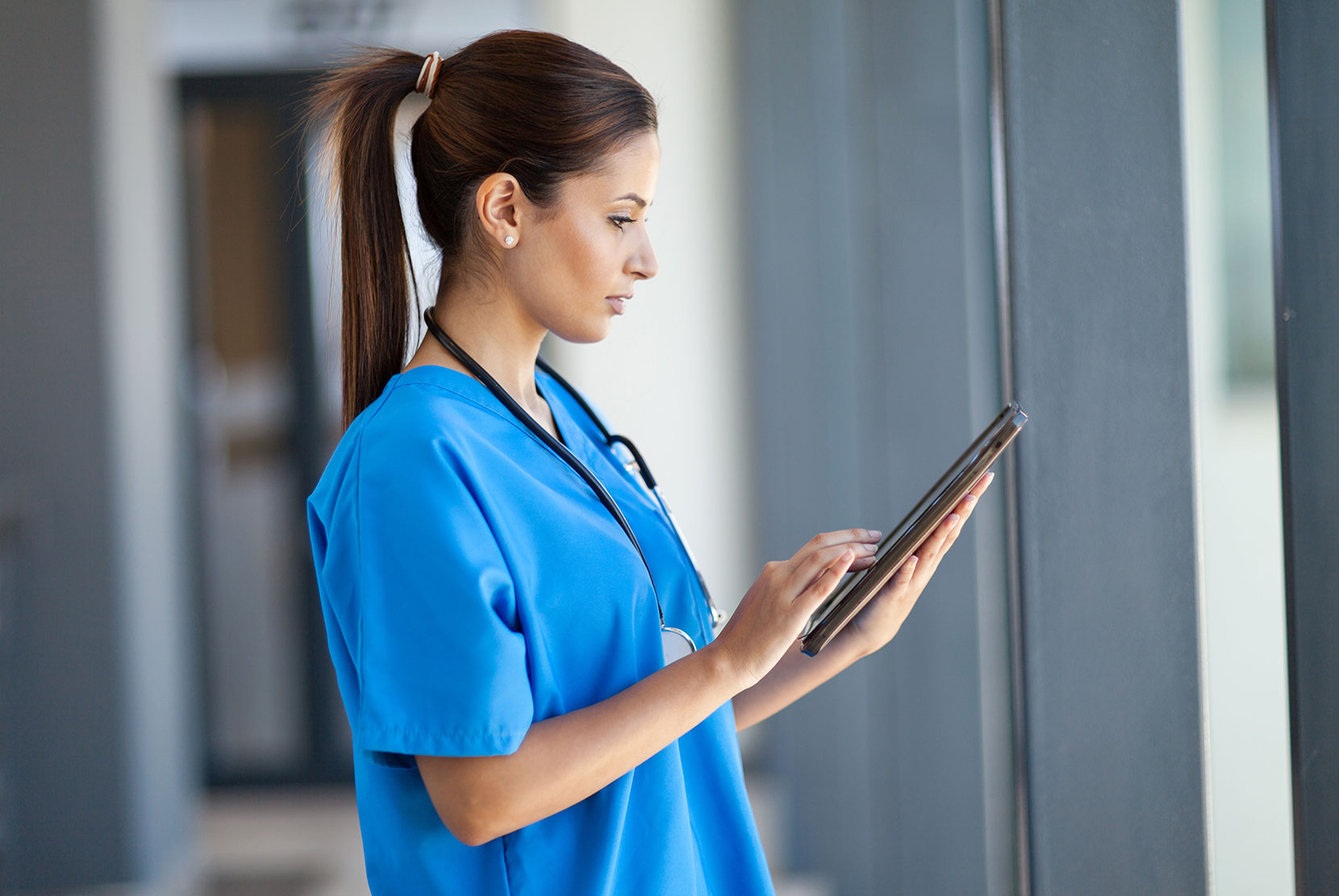 Image of a frontline healthcare worker wearing blue scrubs and a stethoscope and using a mobile tablet in a hospital environment.