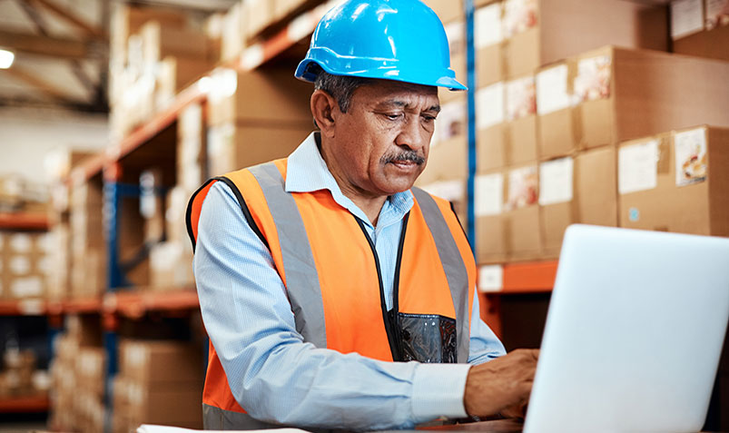 Image shows a frontline worker in a bright blue hard hat and an orange safety vest entering data on a laptop in a warehouse.