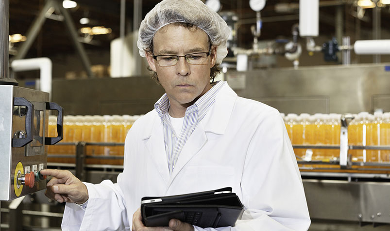 Image shows a factory line operator consulting a mobile tablet while dressed in a white lab coat and wearing a hairnet.