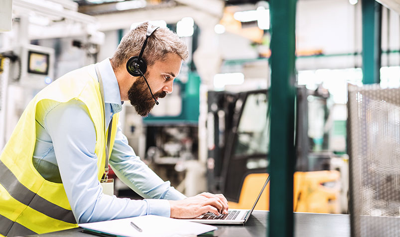 Image shows a frontline worker talking into a mobile headset while typing on a laptop in a warehouse environment.
