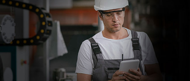 Image shows a frontline worker wearing safety glasses and a grey hard hat, using a mobile tablet within a warehouse