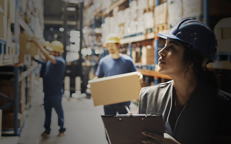 Image of a frontline worker inside of a warehouse wearing a hard hat and taking inventory, while in the background other workers move large cardboard boxes.