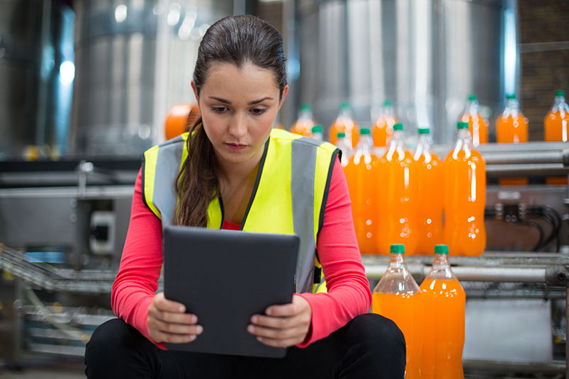 Image shows a frontline worker in factory environment wearing a yellow safety vest and viewing a mobile tablet while sitting in front of many orange soda bottles.